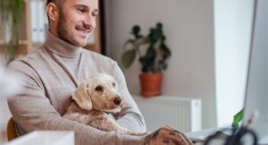 man wearing turtle-neck using computer and smiling with a poodle dog in his lap